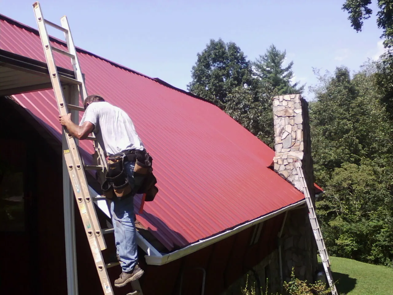 Roofer on a ladder beside a red metal roof and stone chimney — Soffit & Fascia Repair by Roofing Brothers in Attleboro Falls